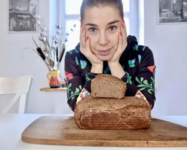 Kobieta z dłońmi podpartymi pod brodą patrzy na pyszny chleb wieloziarnisty. Jeden bochenek leży na drugim, na drewnianej desce.