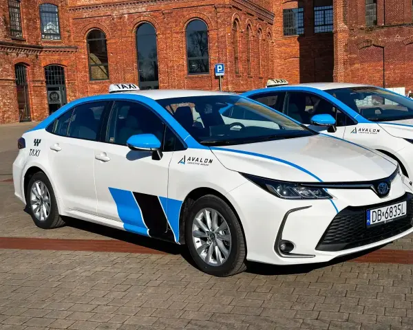 Białe taksówki z niebieskimi pasami i logo Avalon, spełniające uber wymagania samochodu, stoją przed ceglanym budynkiem.