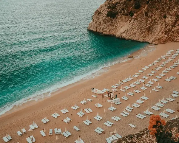 Plaża nad turkusowym morzem tureckim, z rzędami leżaków i skalistym klifem.