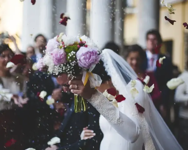 Płatki róż sypią się na młodą parę. W tle widać gości, a na pierwszym planie bukiet panny młodej. To moment, gdy ksiądz na ślubie błogosławi nowożeńców.