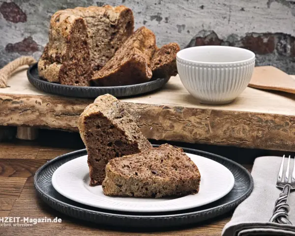 Zwei Stücke eines rustikalen, dunklen Diabetiker Kuchens auf einem Teller, daneben ein ganzer Kuchen mit angeschnittenen Scheiben.