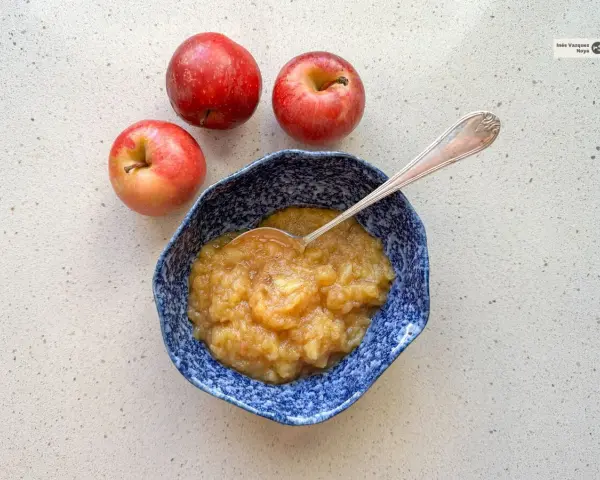 Compota de manzana casera en un cuenco azul, lista para disfrutar. Tres manzanas rojas frescas acompañan este delicioso postre.