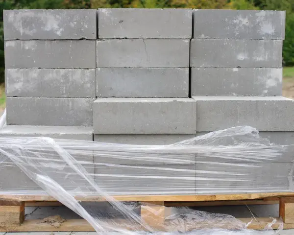 Stos szarych bloczków betonowych na palecie, owiniętych folią. Wymiary bloczków są standardowe, idealne do budowy.