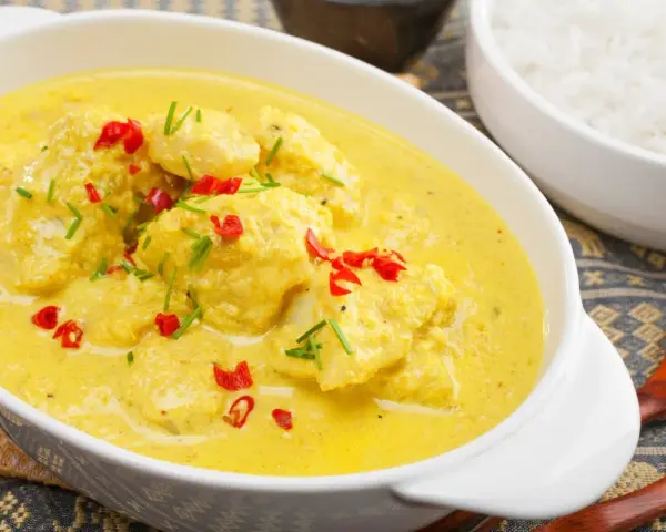 Delizioso pollo al curry con latte di cocco, guarnito con peperoncino rosso e erba cipollina. Accanto, una ciotola di riso bianco.