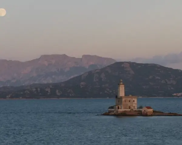 Faro solitario su un'isola sarda, con montagne sullo sfondo e luna piena. Perfetto per itinerari moto Sardegna 7 giorni.