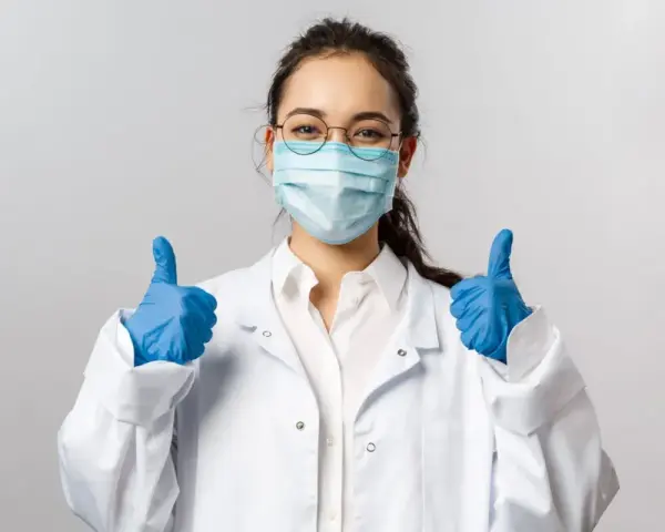 Uśmiechnięta pielęgniarka epidemiologiczna w masce i rękawiczkach pokazuje kciuki w górę.