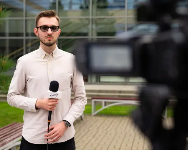 Młody dziennikarz w okularach trzyma mikrofon z logo "FLESZ" przed kamerą.