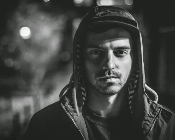 Mroczny portret mężczyzny w kapturze, z zacienioną twarzą i spojrzeniem, które zdaje się mówić o brudnych sercach i trudnych doświadczeniach.