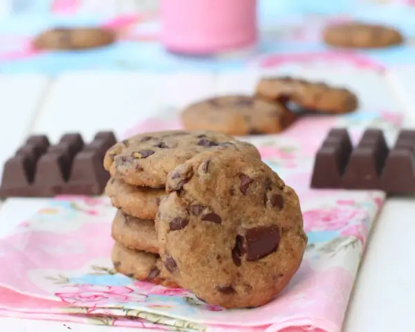 Una pila de galletas de chocolate caseras, con trozos de chocolate visibles, junto a barras de chocolate y un vaso de leche rosa.