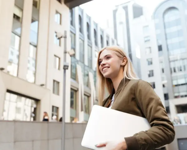 Giovane donna sorridente con laptop, cammina in una città moderna. Lavorare in Svizzera conviene, un futuro brillante ti aspetta.