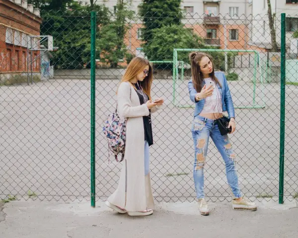 Dwie dziewczyny w modnych stylizacjach, idealnych na outfit do szkoły, stoją przy płocie. Jedna robi sobie selfie, druga przegląda telefon.