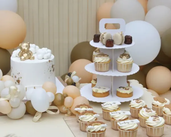 Biały tort z złotym misiem, babeczki i makaroniki na paterze, otoczone balonami w pastelowych kolorach.