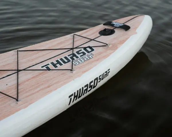 Ein aufblasbares SUP liegt im Wasser. Das Board hat eine Holzoptik und das Logo "THURSO SURF" ist gut sichtbar. Perfekt zum SUP lagern.