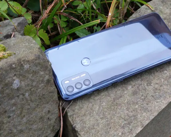 Błyszczący tył telefonu **Moto G 50** z potrójnym aparatem, leżący na kamieniach wśród zieleni.