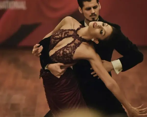 Para tańczy tango, jeden z rodzajów tańca towarzyskiego. Kobieta w bordowej sukni i mężczyzna w czarnym fraku wykonują skomplikowaną figurę.