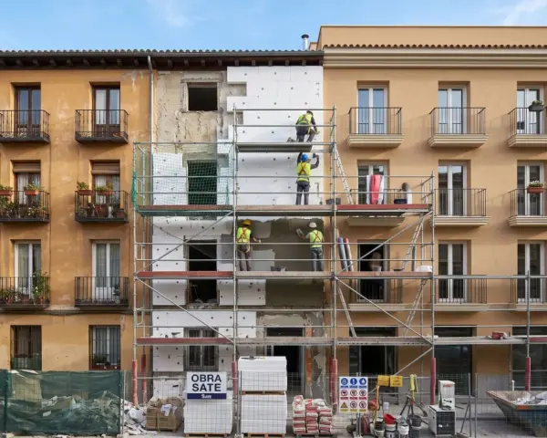 Obreros instalan aislamiento térmico en fachada de edificio. Se ve el **sate lana de roca** en bloques blancos, andamios y materiales de construcción.