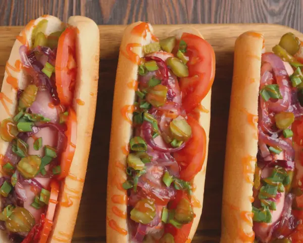 Trzy apetyczne hot dogi z frytkownicy powietrznej, z pomidorami, ogórkami, cebulą i szczypiorkiem, polane sosem.