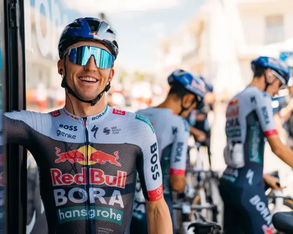 Cyclist smiles on a vuelta rest day, wearing a BORA-hansgrohe kit with Red Bull logos. Other team members and bikes are visible.