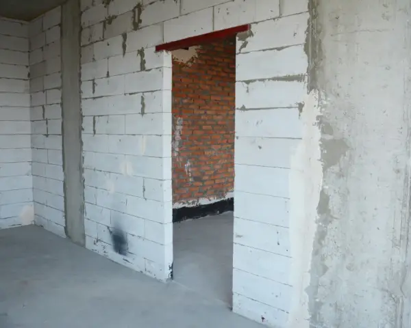 Wnętrze w budowie. Widoczny otwór drzwiowy z czerwonym nadprożem stalowym, ściany z białych pustaków i cegły.