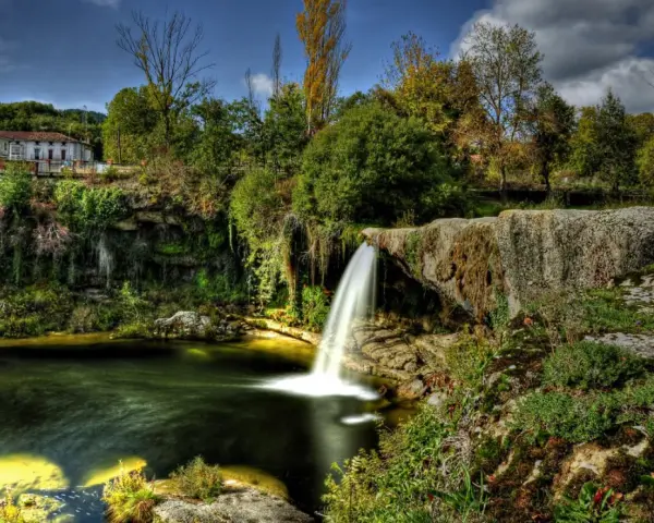 Cascadas en España: una casa rural se asoma entre la vegetación exuberante junto a una cascada que cae en un estanque de aguas verdes.