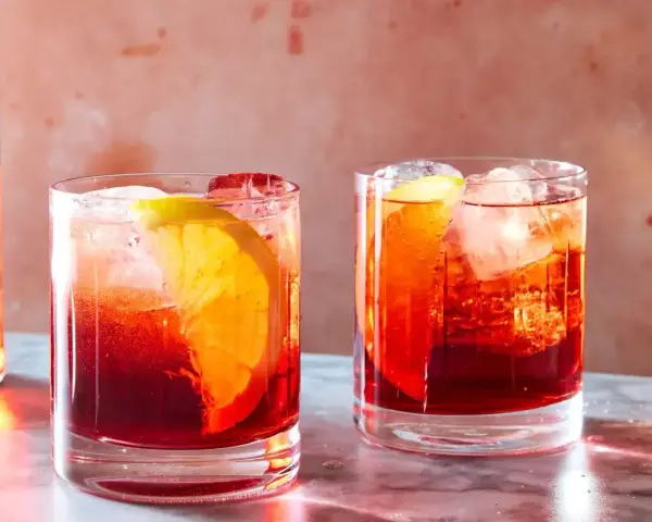 Dwa drinki Negroni z pomarańczą i lodem, obok butelki Campari. Idealny drink negroni przepis na lato.