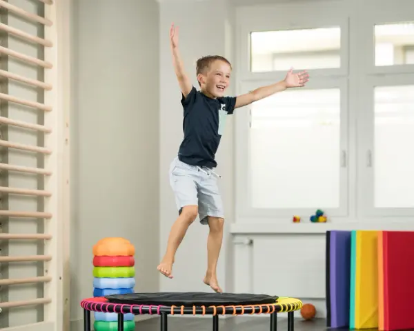 Fröhlicher Junge springt auf Trampolin, Teil der kinder physiotherapie. Kletterwand und bunte Matten im Hintergrund.