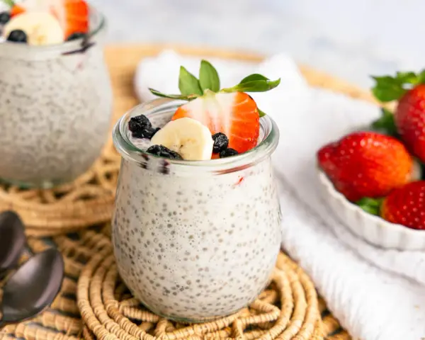 Zwei Gläser mit cremigem Chia Pudding, garniert mit Erdbeeren, Bananen und Blaubeeren. Daneben eine Schale frischer Erdbeeren.