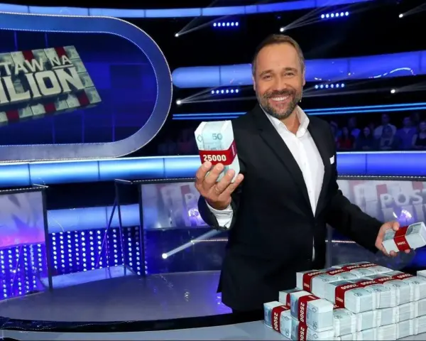 Uśmiechnięty prowadzący teleturniej trzyma w dłoni paczkę pieniędzy. Na stole piętrzą się stosy banknotów.
