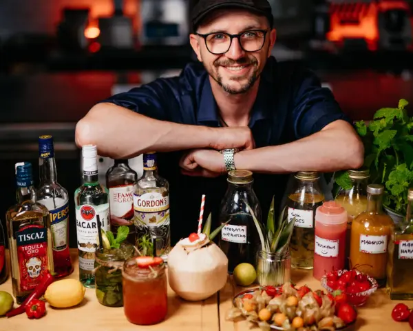 Uśmiechnięty mężczyzna w czapce i okularach za ladą pełną alkoholi i owoców. To przyszłość: drink bar zamiast barmana.