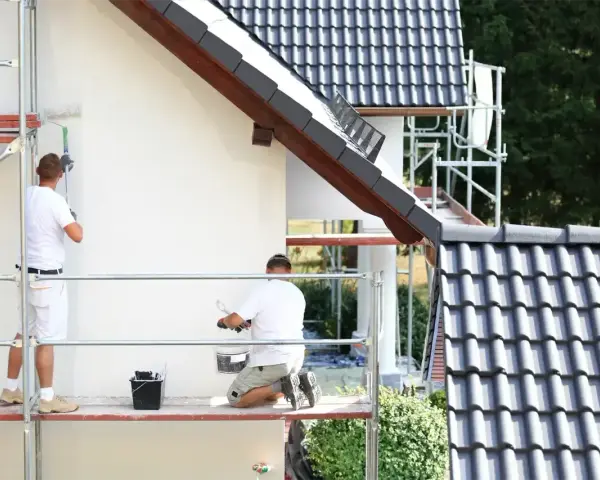 Dwóch mężczyzn na rusztowaniu maluje białą elewację domu. Sprawdź, jaka jest malowanie elewacji cena.