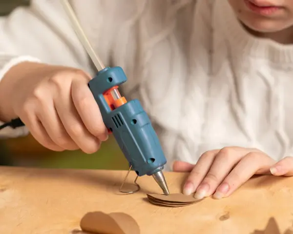 A child uses a hot glue gun to craft heart shapes from brown paper, wondering if hot glue holds up in water for their project.