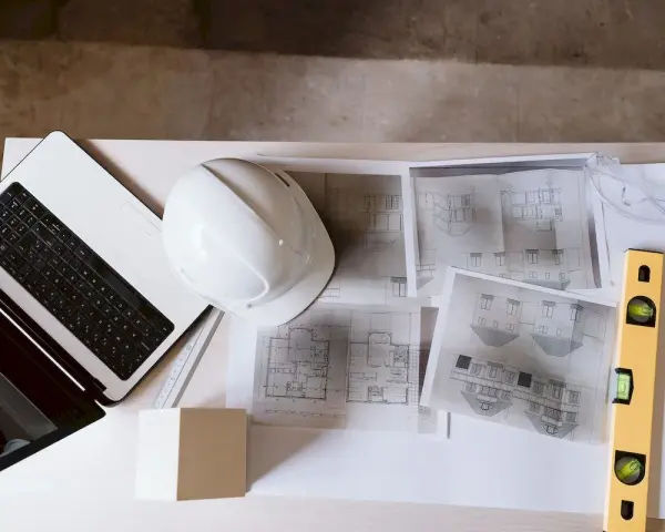 Laptop, kask, poziomica i plany architektoniczne – kompletny zestaw do pracy nad projektem technicznym.