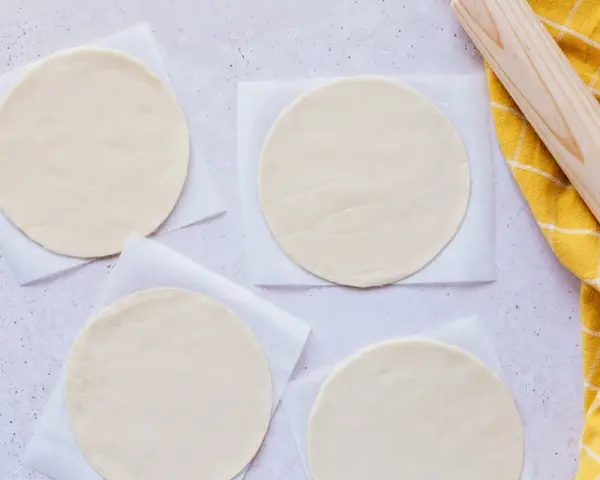 Cuatro discos de masa empanada listos para rellenar, junto a un rodillo y un paño amarillo.