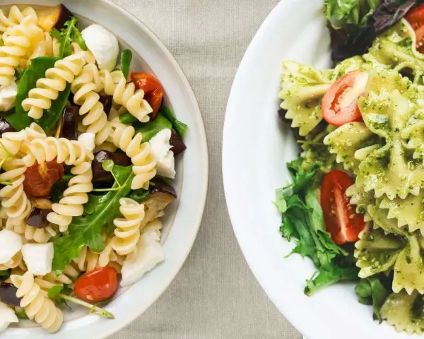 Due ciotole di pasta fredda gourmet: una con fusilli, melanzane, pomodorini e mozzarella; l'altra con farfalle al pesto e pomodorini.
