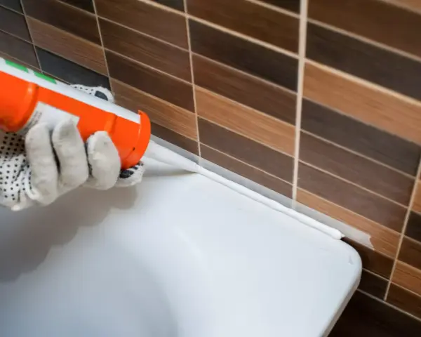 Application d'un joint acrylique ou silicone blanc pour étanchéifier le bord d'une baignoire contre un mur carrelé.