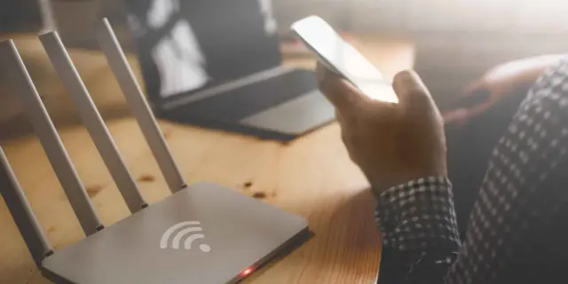 Router Wi-Fi, smartfon w dłoni, laptop na drewnianym biurku.