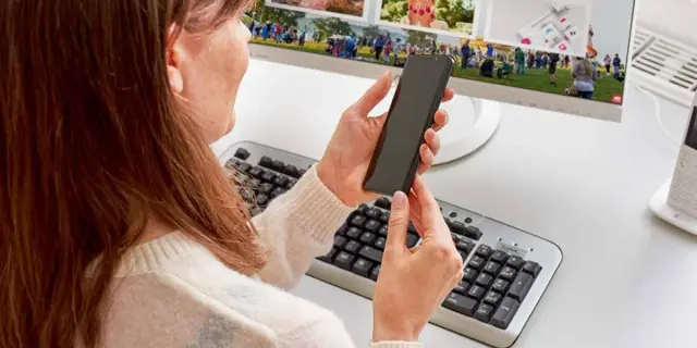 Kobieta trzyma smartfon przed monitorem komputera z klawiaturą i myszką.