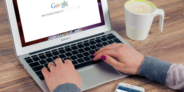 Kobieta pisze na laptopie z otwartą wyszukiwarką Google, obok stoi kubek kawy i smartfon.