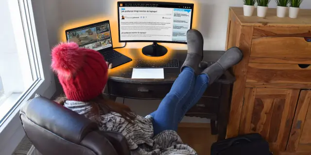 Kobieta w czerwonej czapce i szarych kozakach pracuje przy laptopie i zewnętrznym monitorze.