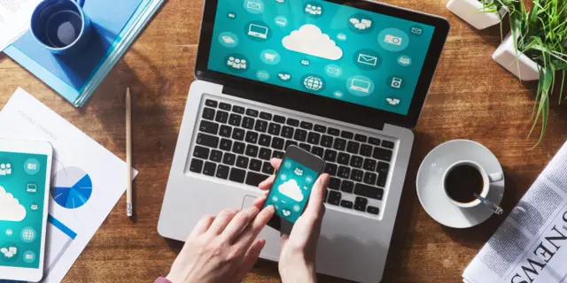 Dłonie trzymające smartfon z ikonami chmury na tle laptopa i dokumentów.