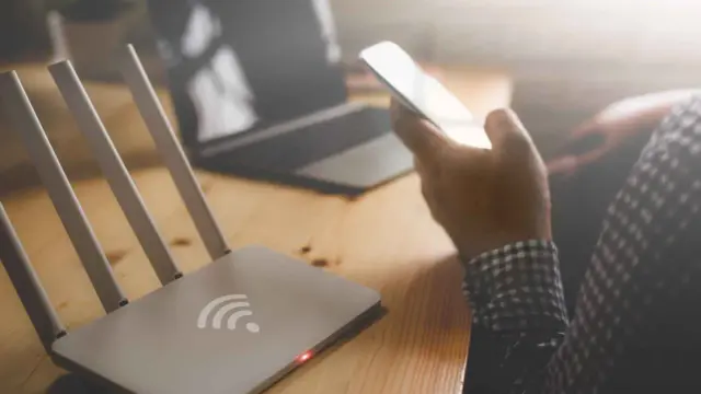 Router Wi-Fi, smartfon w dłoni, laptop na drewnianym biurku.