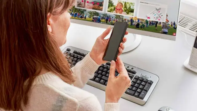 Kobieta trzyma smartfon przed monitorem komputera z klawiaturą i myszką.