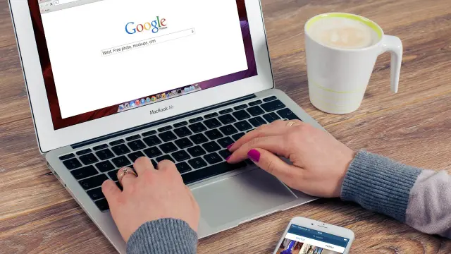 Kobieta pisze na laptopie z otwartą wyszukiwarką Google, obok stoi kubek kawy i smartfon.