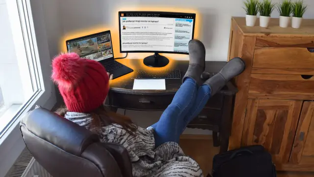 Kobieta w czerwonej czapce i szarych kozakach pracuje przy laptopie i zewnętrznym monitorze.
