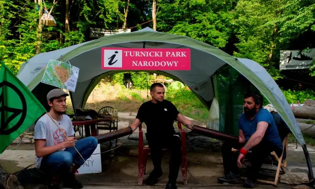 Trzech mężczyzn siedzi pod namiotem z napisem "Turnicki Park Narodowy". Obok zielona flaga z symbolem.