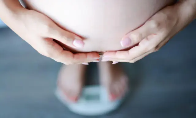 Dłonie obejmujące brzuch ciężarnej, stojącej na wadze. Schudłam w ciąży 10 kg, co widać na wadze.