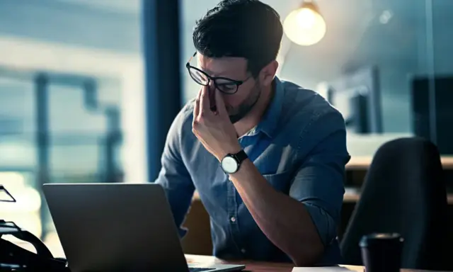 Mann reibt sich erschöpft die Augen vor dem Laptop. Ein Hackerangriff auf Privatpersonen kann zu solchen Belastungen führen.