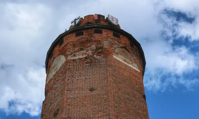 Wieża zamku krzyżackiego w Brodnicy, z cegły, z herbami na szczycie, na tle błękitnego nieba z chmurami.
