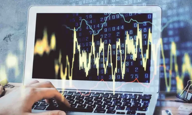 Dłoń wpisuje kod na klawiaturze laptopa, na ekranie wykresy i liczby symbolizujące handel algorytmiczny.