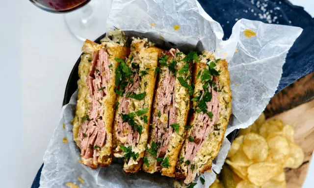 En saftig reuben sandwich, skuren i fyra bitar och toppad med färsk persilja. Serveras med krispiga potatischips och en glas rödvin.